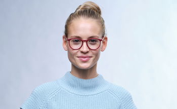 Das Bild zeigt eine Frau mit einer roten Brille und einem hellblauen Pullunder.